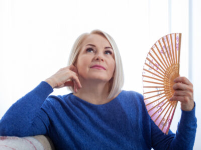 A woman in her fifties sits and is fanning herself with a handheld fan, her face flush with the heat of menopause.