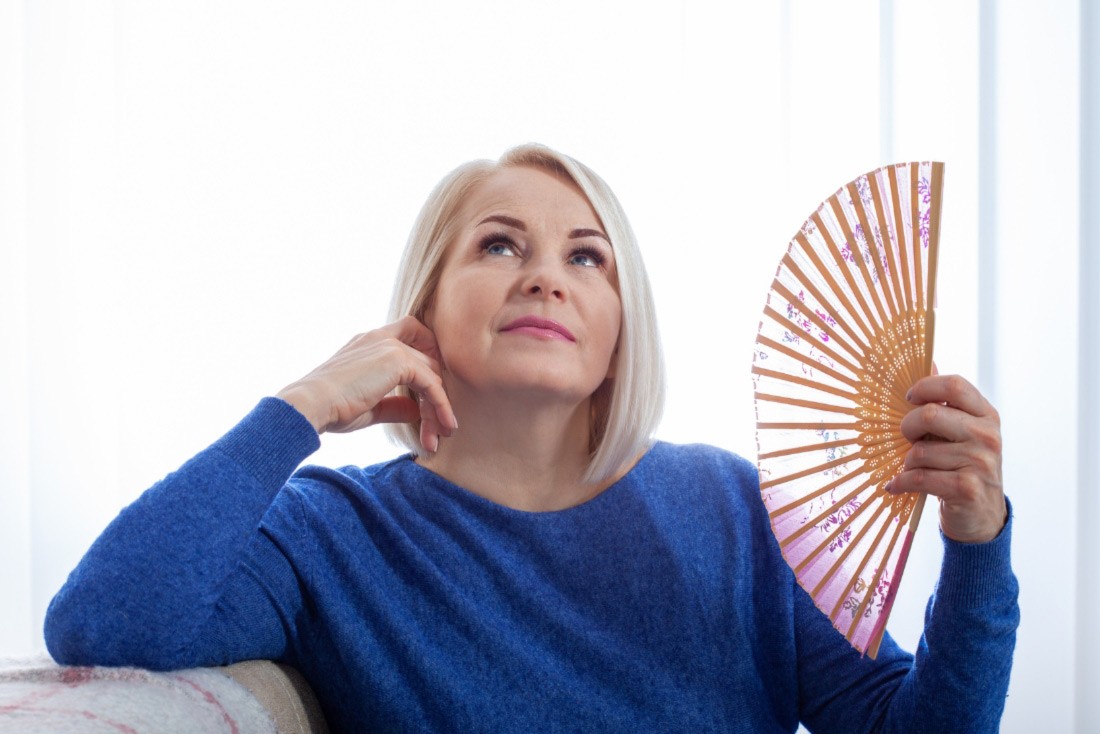 A woman in her fifties sits and is fanning herself with a handheld fan, her face flush with the heat of menopause.