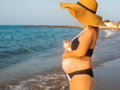 A pregnant woman wearing a wide brimmed hat and black bikini stands on a beach and uses sunscreen.