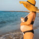 A pregnant woman wearing a wide brimmed hat and black bikini stands on a beach and uses sunscreen.