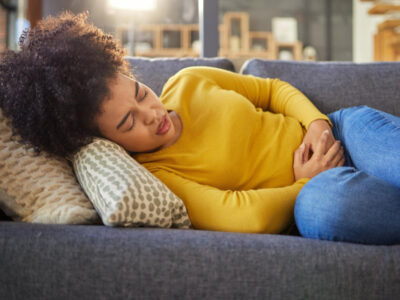 A woman lies on a couch holding her lower abdomen, appearing to experience pelvic pain or discomfort. She is wearing a yellow sweater and blue jeans. The image illustrates common symptoms of PCOS such as abdominal pain or cramping.
