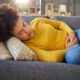 A woman lies on a couch holding her lower abdomen, appearing to experience pelvic pain or discomfort. She is wearing a yellow sweater and blue jeans. The image illustrates common symptoms of PCOS such as abdominal pain or cramping.