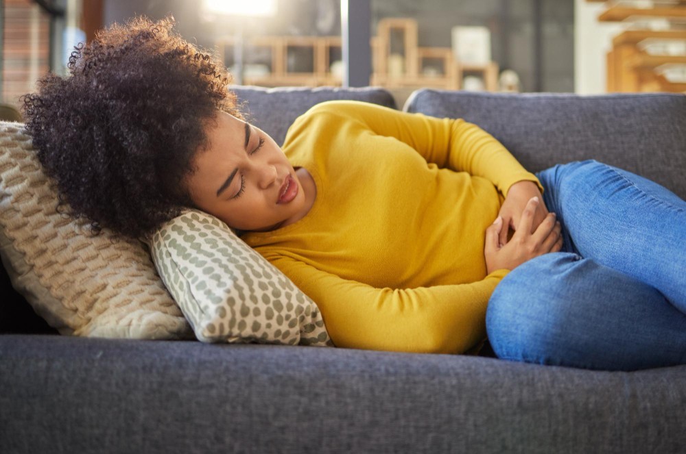 A woman lies on a couch holding her lower abdomen, appearing to experience pelvic pain or discomfort. She is wearing a yellow sweater and blue jeans. The image illustrates common symptoms of PCOS such as abdominal pain or cramping.
